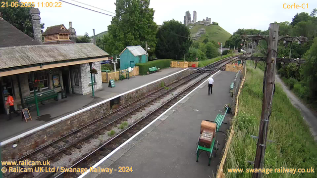A small train station platform with a stone building on the left, featuring green benches, flower baskets, and a person in a bright orange vest. Two other people are on the platform; one walking away from the camera and another standing near the building. Railroad tracks run through the center, with a grassy area and trees on the right. In the background, a green hill with ruins or castle remains perched atop it. A wooden telephone pole and some power lines are visible on the right side of the image. The scene appears to be during the daytime on a cloudy day.