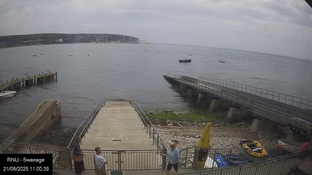 A view of a small harbor with a concrete slipway leading into the calm sea. There are three people standing near the entrance to the slipway, talking. To the right, there is a yellow life raft or boat and some equipment. In the water, several boats are anchored, and a large headland with cliffs is visible in the distance under a cloudy sky.