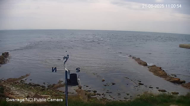 A view of a calm ocean with a rocky shoreline in the foreground. The sky is partly cloudy with some light clouds. There is a weather vane and anemometer mounted near the bottom left corner, and the timestamp shows June 21, 2025, at 11:00:54. The image is labeled as from the Swannage NCI Public Camera.