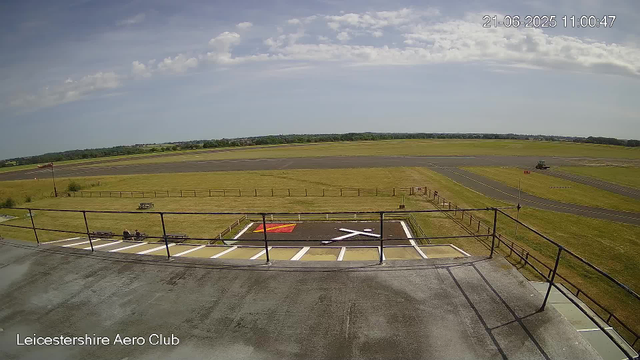 A view from a webcam at Leicestershire Aero Club shows a small aircraft parking area with marked spaces, a grassy field, and a runway in the background. The sky is partly cloudy with scattered clouds, and the scene is well-lit by sunlight. The timestamp on the image reads 21-06-2025 11:00:47.