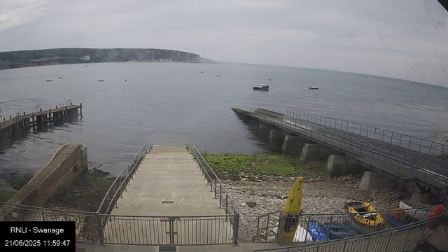 A view of a small pier extending into calm water, with a boat docked nearby. There are two breakwaters with railings, one on each side, and a concrete ramp or walkway leading from the shore into the water. In the foreground, there is a railing enclosing a small area with a yellow life-saving float and some equipment. The sky is overcast with grey clouds, and in the distance, there are white cliffs along the coastline.