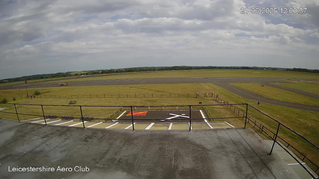 A view from a webcam capturing an airfield at the Leicestershire Aero Club on a partly cloudy day. In the foreground, there's a parking area with labeled lines and a red and white helipad symbol. Fenced grass surrounds the parking area. Beyond the grass, a runway extends across the image with a small vehicle or aircraft on it. In the distance, a person stands near the fence, and the landscape stretches out with open fields and a few scattered trees under a partly cloudy sky. The date and time are displayed in the top right corner as June 21, 2025, at 12:00:37.