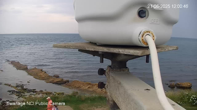 A webcam captures a view of a seaside landscape with a cloudy sky. In the foreground, there is a white device, possibly a weather instrument or camera, mounted on a wooden and metal structure. A white cable extends from the device. At the bottom left corner, a person wearing a pink jacket and a white hat is sitting or standing near the grassy area, with rocks and the water's edge nearby. The date and time displayed on the image are June 21, 2025, at 12:00:46. The footage is credited to Swannage NCI Public Camera.