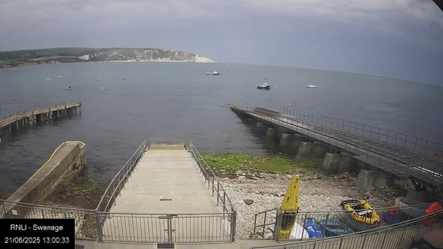 A view of a small harbor with boats on calm water, a stone and concrete pier extending into the sea, and a rocky shoreline with some green moss. In the background, there are cliffs and land formations under a cloudy sky. A yellow warning sign and some equipment are visible near the edge of the shoreline.