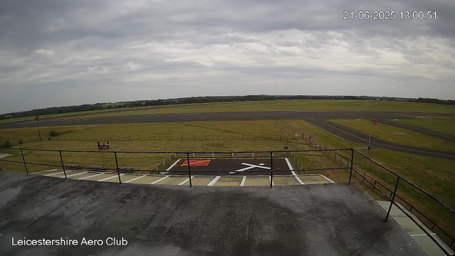An empty tarmac area with a helicopter landing pad marked by a white hazard symbol and a red square. There is a railing in the foreground, and a bench with two people sitting on it on the grass. Beyond the area, there is open grassland and a runway, with a cloudy sky overhead. The timestamp reads "21-06-2025 13:00:51" and the location is marked as "Leicestershire Aero Club."