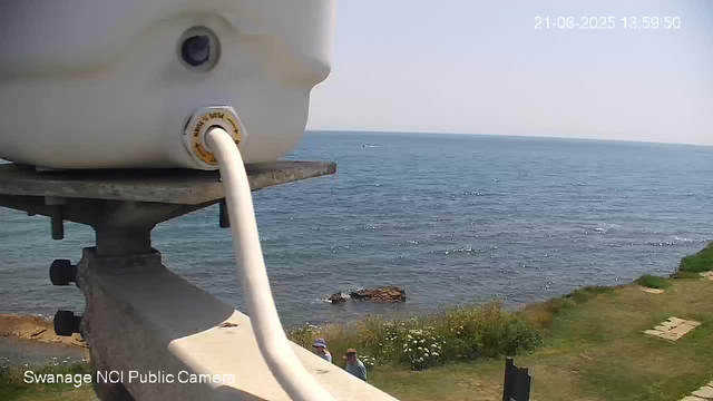 A webcam captures a view of the ocean with gentle waves and a small boat in the distance. In the foreground, part of the camera equipment and a railing are visible, with a grassy area and some people standing near the shore. The timestamp in the top right corner indicates it was taken on June 21, 2025, at 13:59:50.
