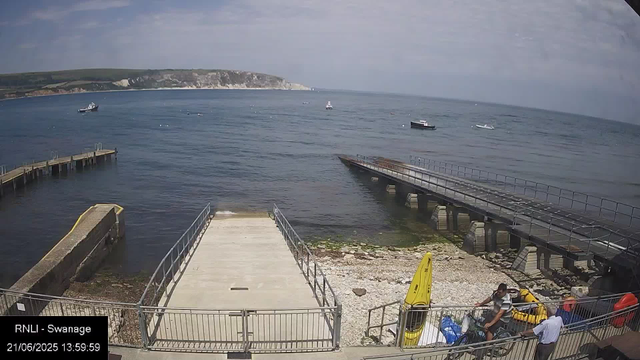 A view of a seaside area with a pier extending into the water, and several boats floating in the ocean. In the foreground, there are two people near yellow rescue equipment and a person on a bicycle. The background shows a hilly coastline under a partly cloudy sky.