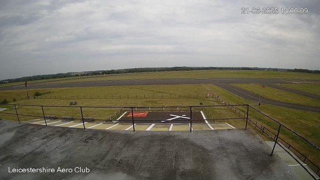 A view from a webcam at Leicesterhire Aero Club showing a clear, open outdoor area with a tarmac runway extending into the distance. The sky is partly cloudy, and there are some distant trees and low-lying buildings on the horizon. In the foreground, there is a small fenced platform with a marked helipad. The timestamp reads 21-06-2025 14:00:09.