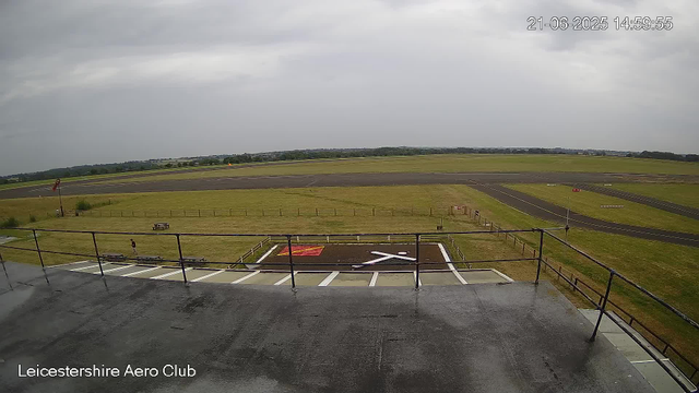 The image shows a view from a webcam at Leicesterhire Aero Club. In the foreground, there is a concrete viewing platform with a metal railing, a red and white helicopter landing pad marked with a helicopter symbol, and a few empty benches. Beyond the platform, the grassy area stretches out with a small flagpole on the left and a person walking nearby. In the background, there is a paved runway with multiple taxiways and open fields under a cloudy sky. The timestamp on the top right corner indicates it was taken on June 21, 2025, at 14:59:55.