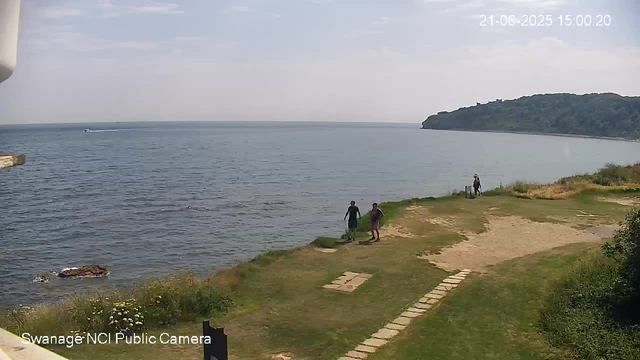 A view of a coastal scene with a calm sea extending to the horizon, where a boat is leaving a wake. On the right side, there is a large green hillside. In the foreground, three people are walking along a grassy and sandy shoreline; two are closer together near the water, and one is farther away near the edge of the land. The sky is partly cloudy with light gray and white clouds. The timestamp in the top right corner shows "21-06-2025 15:00:20," and the bottom left corner indicates "Swanage'NCI Public Camera."