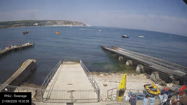 A view of a seaside area with boats on the water and a concrete pier extending into the sea. There are several boats floating on the water, and a tall chalk cliff can be seen in the background under a partly cloudy sky. In the foreground, there is a fenced area with people standing and watching, along with some yellow and orange water safety equipment and small vehicles. The timestamp on the image is "21/06/2025 15:00:55."