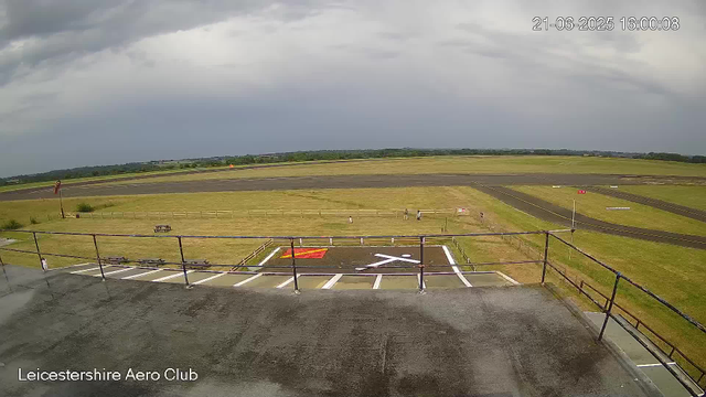 A wide view of an airfield showing a runway in the distance under a cloudy sky. In the foreground, there is a concrete platform with a safety railing, a marked helipad with a red and white circle, and some parking spaces. A few people are walking on the grass and near the helipad. The landscape beyond the airfield extends to a horizon of trees and open fields. The timestamp in the top right corner reads "21-06-2025 16:00:08." The text "Leicestershire Aero Club" is at the bottom left corner of the image.