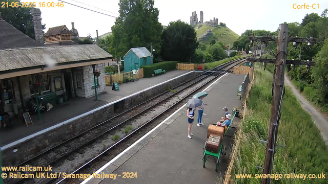 A small train station platform with three people standing and sitting. One person is holding an umbrella, while others are seated on benches. There are a few potted plants, a wooden chest, and a green cart with some items on it. Behind them, train tracks run parallel, with a yellow fence along the side. On the left, there's a building with a stone exterior and hanging flower baskets. In the background, a hill with a castle ruins structure is visible, along with green trees and a lamppost. An overhead power line runs across the scene, and a narrow footpath leads away to the right. The timestamp indicates the photo was taken on June 21, 2025, at 4:00 PM.