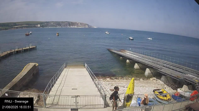 A view of a small harbor on a sunny day with calm water and several boats anchored in the distance. There is a concrete pier extending into the water, and a floating dock is connected to the shoreline. Two people are walking near the dock, one carrying a bag, and there are yellow and red jet skis or small boats parked on the shore. The background features a distant coastline with white cliffs and green hills under a partly cloudy sky. The bottom left corner has a timestamp and location label reading "RNLI - Swanage 21/06/2025 16:00:51."