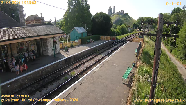 A small train station platform with a stone building on the left side, where several people are sitting and standing. There are parked benches along the platform, a small blue shed behind a green hedge, and a row of flowers in pots on a green bench to the right. In the background, a lush green hillside features a castle ruin at the top, with some trees and power lines. The sky is clear and bright, indicating a sunny day. There are train tracks running parallel to the platform, extending into the distance toward the right side of the image.