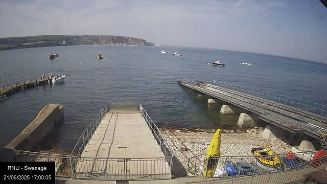 A scenic view of a small harbor with calm water, several boats floating in the distance, and a rocky coastline with cliffs in the background. There are two concrete docks extending into the water, one with a person near a small boat tied to it. In the foreground, there is a fenced area with a yellow kayak, some small vehicles, and other equipment; a yellow coned sign is also visible. The sky is partly cloudy with patches of blue.