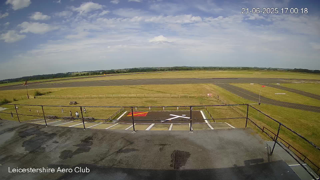 A view from a webcam at Leicestershire Aero Club showing a clear sky with scattered clouds, a runway in the distance, and a grassy area with several small groups of people near a fenced lookout point. The timestamp indicates it is June 21, 2025, at 5:00 PM.