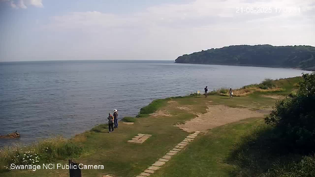 A scenic view of a coastal area shows a grassy and sandy shoreline with a few people walking and standing near the water. In the background, there is a large hill or small mountain covered in greenery, extending out into the sea. The sky is partly cloudy, and the ocean appears calm. The image is from a public webcam, as indicated by the text at the bottom left corner.