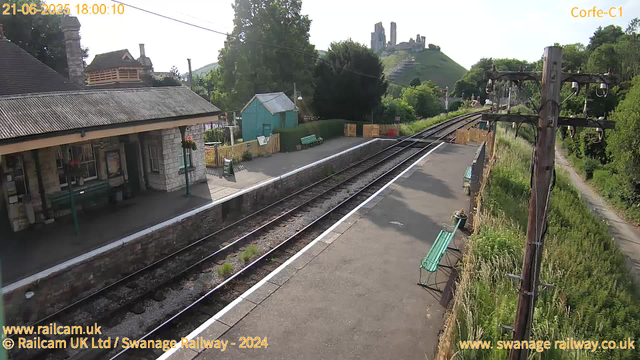A small railway station platform with a stone building on the left, featuring a roof with tiles and a chimney. There are benches, flower pots, and signs along the platform. Railway tracks run through the center, curving slightly into the distance. To the right, there is an old wooden utility pole with electrical wires and insulators, and a narrow path with greenery and bushes beside it. In the background, a lush green hillside with a castle ruin sits atop, overlooking the scene. The sky is clear and bright, indicating a sunny day. The timestamp on the image reads "21-06-2025 18:00:10," and the URL text is visible at the bottom left and right.