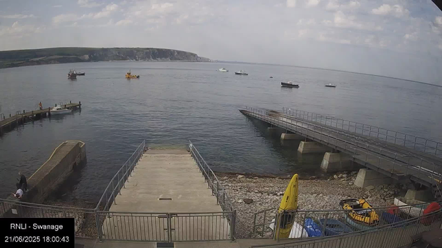 A view from a webcam showing a marina with a concrete dock extending into calm water. Several small boats are anchored in the bay, with a hill or cliff in the background. The sky is partly cloudy, and a person is standing on the pier near a small boat. There are some safety equipment and vehicles near the shoreline, and a yellow caution sign is visible on the right side of the foreground. The timestamp indicates the date as 21/06/2025 at 18:00:43, and the location as RNLI - Swanage.