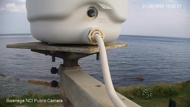 A close-up view of a white electronic device mounted on a concrete lookout, with a cable connected to it, overlooking a body of water with gentle waves. The background shows the horizon and a cloudy sky. The timestamp in the top right corner reads “21-06-2025 18:00:51,” and the bottom left indicates it is from the “Swanage NCI Public Camera.”