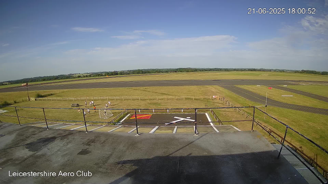 An outdoor scene at Leicesterhire Aero Club showing a clear sky with a few clouds, a grass field with a small group of children playing, a runway in the background, and parked motorcycles in the foreground. The image includes a timestamp of June 21, 2025, at 18:00:52.