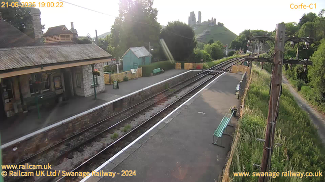 A small railway station with a stone building on the left, a platform with a green bench, a small turquoise shed, and a wooden bus stop sign. Tracks run straight through the scene, with some grass growing between the rails. Wooden fencing separates parts of the station. To the right, there are tall grassy plants and a weathered wooden telephone pole with wires extending across the scene. In the background, there is a lush green hillside with the ruins of a castle on top, and the sky is clear with the sun shining brightly. The timestamp indicates it is early evening.