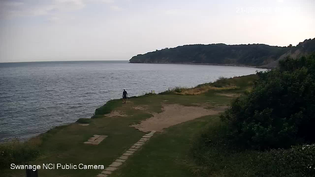A person is walking along a grassy and sandy path near the edge of a body of water, with a large wooded hill in the background. The sky is overcast, and the time stamp shows the date and time as 21:06:23. The image is from a Swannage NCI public camera.