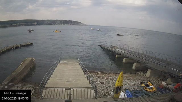 A view of a harbor with a concrete boat ramp extending into the water in the foreground. To the right of the ramp, there is a yellow caution wet floor sign and some equipment, including a yellow inflatable boat. Several boats are anchored in the water, varying in size and color. In the background, there is a distant coastline with cliffs under a cloudy sky. The image is timestamped "21/06/2025 19:00:35" and labeled "RNLI - Swanage".