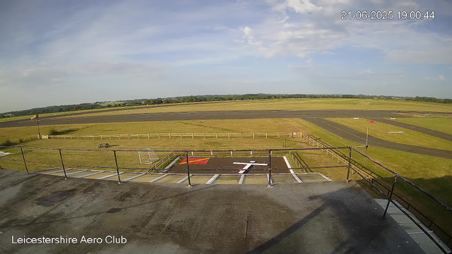 An empty rooftop observation area with a railing overlooking a wide outdoor airfield or airport runway, with grassy fields and a partly cloudy sky in the background. The image timestamp shows "21-06-2025 19:00:44" and the location is labeled as "Leicestershire Aero Club."