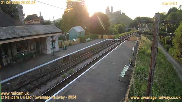 A small railway station platform at sunset, with two empty benches along the edge. A wooden building with a corrugated metal roof and flower baskets hanging outside is on the left. Railway tracks run through the center, curving slightly to the right in the distance. A telegraph pole with wires is on the right side, and a narrow footpath runs parallel to the tracks, bordered by grass and bushes. In the background, there is a hill with castle ruins on top, and trees surrounding the area. The sun is low in the sky, casting a bright glow and lens flare.