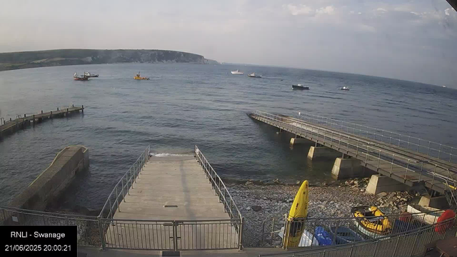 A view of a seaside dock area with a concrete pier extending into the water. Several boats are anchored in the distance on the calm sea, and a headland with cliffs can be seen on the horizon. In the foreground, there are yellow rescue equipment and a small shoreline with rocks. The scene appears to be during early evening, with partly cloudy skies.