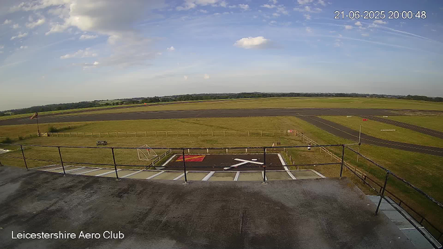 A wide view of Leicestershire Aero Club's airfield shows a clear sky with some scattered clouds, a grassy area with a small fenced section, and a paved helipad with white markings and a red section. In the distance, there are runways and open fields. The image is timestamped "21-06-2025 20:00:48" in the top right corner. The environment appears calm and open.