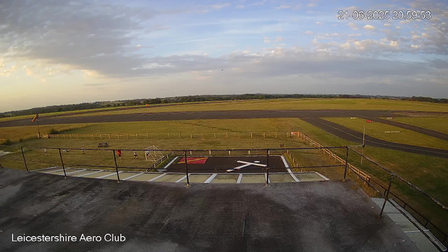 An overview of an airfield runway with a small grassy area in the foreground where a few people are walking. There is a helipad with a black and white "H" in the foreground, and a red and white markings area. The runway stretches into the distance under a partly cloudy sky, with a few small trees and fields visible beyond the airfield perimeter. The timestamp in the upper right corner reads "21-06-2025 20:59:53," and the bottom left corner has the text "Leicestershire Aero Club."