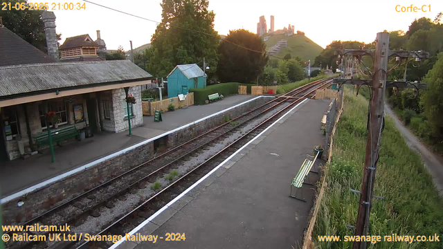 A small railway station platform at sunset with benches along the side, a stone building with hanging flower baskets, a small blue shed, and a fence with potted plants. Railway tracks run parallel to the platform, which is lined with white paint. Utility poles with wires run along the edge of the platform, and there's a dirt path with grass and wildflowers on the right side. In the background, there are trees, a hill with a historic castle or ruins on top, and a sky with warm sunset light.