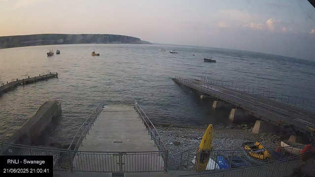 A view of a harbor with calm water and several boats floating in the distance. There are two concrete piers extending into the water, one on the left and one on the right. On the pebble beach in the foreground, there are yellow safety signs and some equipment, including a kayak and a small vehicle. A coastal cliff with greenery is visible in the background under a partly cloudy sky during sunset.