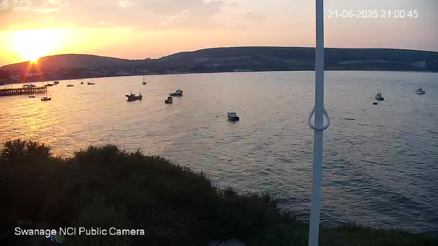 A scenic view of a calm bay at sunset with several boats anchored on the water. The sun is low on the horizon, casting a warm glow and reflecting off the surface of the bay. In the background, there are rolling hills under a partly cloudy sky. A white pole is in the foreground on the right side, and a small pier extends into the water on the left side. The timestamp in the top right corner indicates the date and time as June 21, 2025, at 9:00:45 PM. The bottom left corner displays the label "Swanage NCI Public Camera."