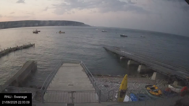 A view of a waterfront scene at dusk, showing a concrete pier extending into the water with metal railings, several boats floating on the calm sea, a hilly coastline in the background under a cloudy sky, and some safety equipment and vehicles on the shore in the foreground.