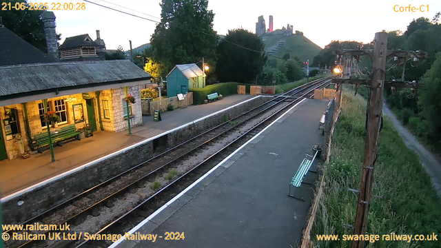 A small train station platform at sunset, with a stone building on the left displaying green benches and hanging flower baskets. There is a narrow railway track running alongside the platform, with a wooden fence and some vegetation on the right. In the background, there are trees, a small teal-colored shed, and a grassy hill with a castle ruin at the top. The sky is faintly illuminated, indicating early evening. The image has a timestamp indicating June 21, 2025, at 10:00 PM.