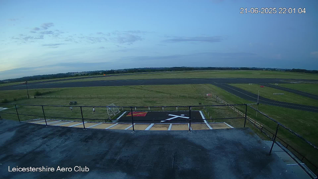 An empty Helipad with a white helicopter symbol and red square marking, located on a rooftop overlooking a grassy airfield with runways and a few small people walking on the grass. The sky is partly cloudy with a mix of blue and some clouds, and the timestamp reads 21-06-2025 22:01:04. The bottom left corner has the text "Leicestershire Aero Club."