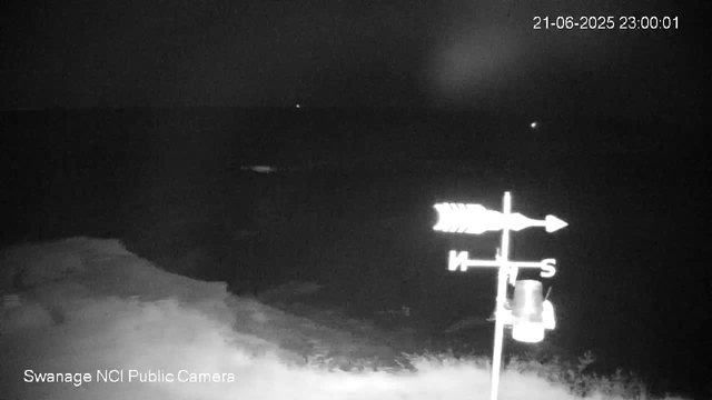 A black-and-white night-time image from a webcam shows a wooden signpost with an arrow pointing to the right, labeled with the cardinal directions North (N), South (S), East (E), and West (W). The signpost is situated on a road or pathway with some terrain, possibly sand or gravel, visible at the bottom left. The sky is dark with a few faint stars visible. The timestamp in the top right corner indicates the date as June 21, 2025, and the time as 23:00:01. The bottom left corner has the text "Swanage NCI Public Camera."