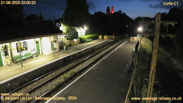 A nighttime image of a small train station platform with a building on the left illuminated by yellowish lights. The platform has benches, flower baskets, and a green sign that reads "WAY OUT." The railway tracks run between the platform and a narrow path on the right, which is separated by a fence and a wooden utility pole with wires. In the background, there are trees, a hill, and a structure on top illuminated with red lights. The sky is dark with some faint clouds visible. The timestamp on the top left shows "21-06-2025 23:00:22," and the top right indicates "Corfe-C1." The bottom of the image contains credits for the railway company and website.