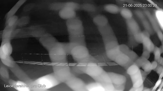 A black and white image from a webcam shows a blurred scene with circular bokeh effects. The timestamp on the image reads "21-06-2025 23:00:29," and there is a fence or railing visible at the bottom of the frame. The text "Leicestershire Aero Club" is located in the lower left corner. The overall scene appears to be taken at night or in low light conditions.