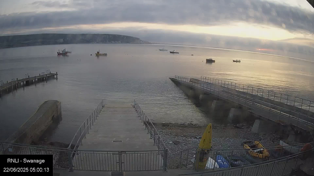 A view of a calm harbor at sunrise with several small boats floating on the water. There are two piers extending into the water, one on the left and one on the right, and a pathway with railings leading into the water in the foreground. The sky is partly cloudy with the sun just beginning to rise, casting a soft glow over the scene. Some equipment, including a yellow lifeboat and a yellow caution sign, is visible near the waterfront.