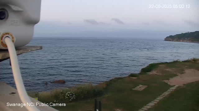 A view of a coastal landscape with a body of water stretching into the distance, a rocky shoreline on the right, and some grassy areas with a paved path in the foreground. The sky is overcast with gray clouds. A camera is partly visible on the left side of the image. The date and time are displayed in the top right corner as "22-06-2025 05:00:12."
