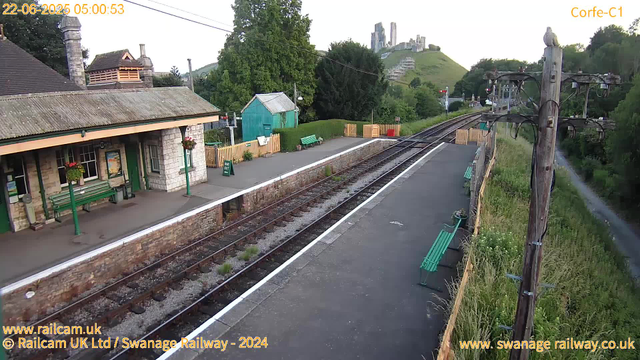 A small railway station platform with a stone building on the left, decorated with hanging flower baskets. There are several green benches along the platform, some with potted plants nearby. Electric poles with wires run alongside the tracks on the right. In the background, lush green trees, a small turquoise shed, and a hill with an ancient castle ruin atop it are visible. The scene appears to be early morning, with the date and time displayed as June 22, 2025, at 05:00:53.