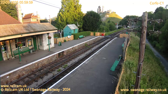 A small train station platform with a building on the left that has a stone facade, green benches, and hanging flower baskets. There are railway tracks running through the middle, with a signal in the distance showing red. To the right, there are wooden utility poles with wires, green benches, and grass overgrown alongside the platform. In the background, a lush green hillside with a castle ruin on top is visible, and the sky is clear with bright sunlight illuminating the scene. The timestamp indicates it is early morning, June 22, 2025.