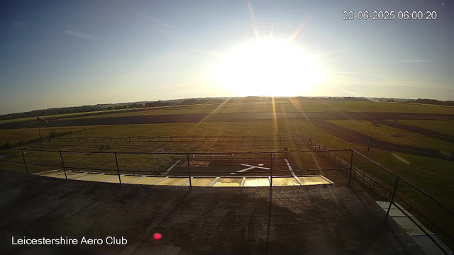 A view from a webcam at Leicestershire Aero Club shows a bright, sunny morning with the sun low on the horizon, casting long rays across open grassy fields and a runway. The landscape is flat with distant trees and clear skies. The timestamp indicates the date and time as June 22, 2025, at 6:00 AM. The foreground includes a rooftop with railings and some small structures.