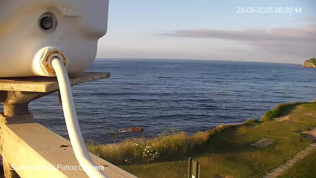 A webcam captures a peaceful coastal scene during what appears to be early morning or late evening. The image shows a calm ocean with gentle waves extending to the horizon. To the right, there is a small section of land with some greenery and a few trees. The sky is partly cloudy with a mix of blue and soft pinkish hues. In the foreground on the left side, part of the webcam device and its power cable are visible, with a portion of a wooden railing beneath it. The timestamp on the image reads 22-06-2025 06:00:44.