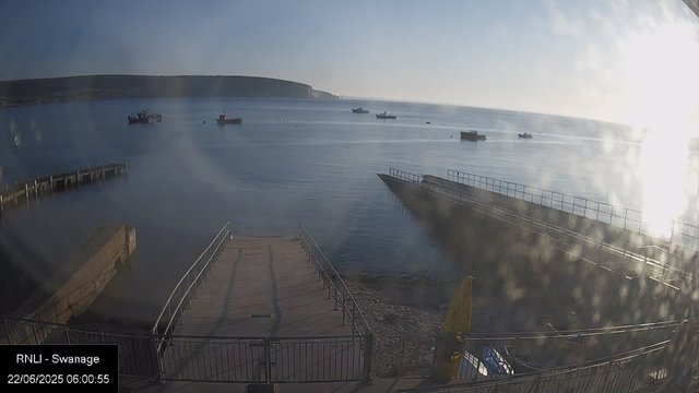 A calm seaside scene at dawn with a clear sky. Several boats are anchored in the water, and a small pier extends into the sea on the left side. In the background, there are rolling hills or cliffs along the horizon. The water appears smooth, reflecting the early sunlight, and the overall atmosphere is peaceful and quiet.